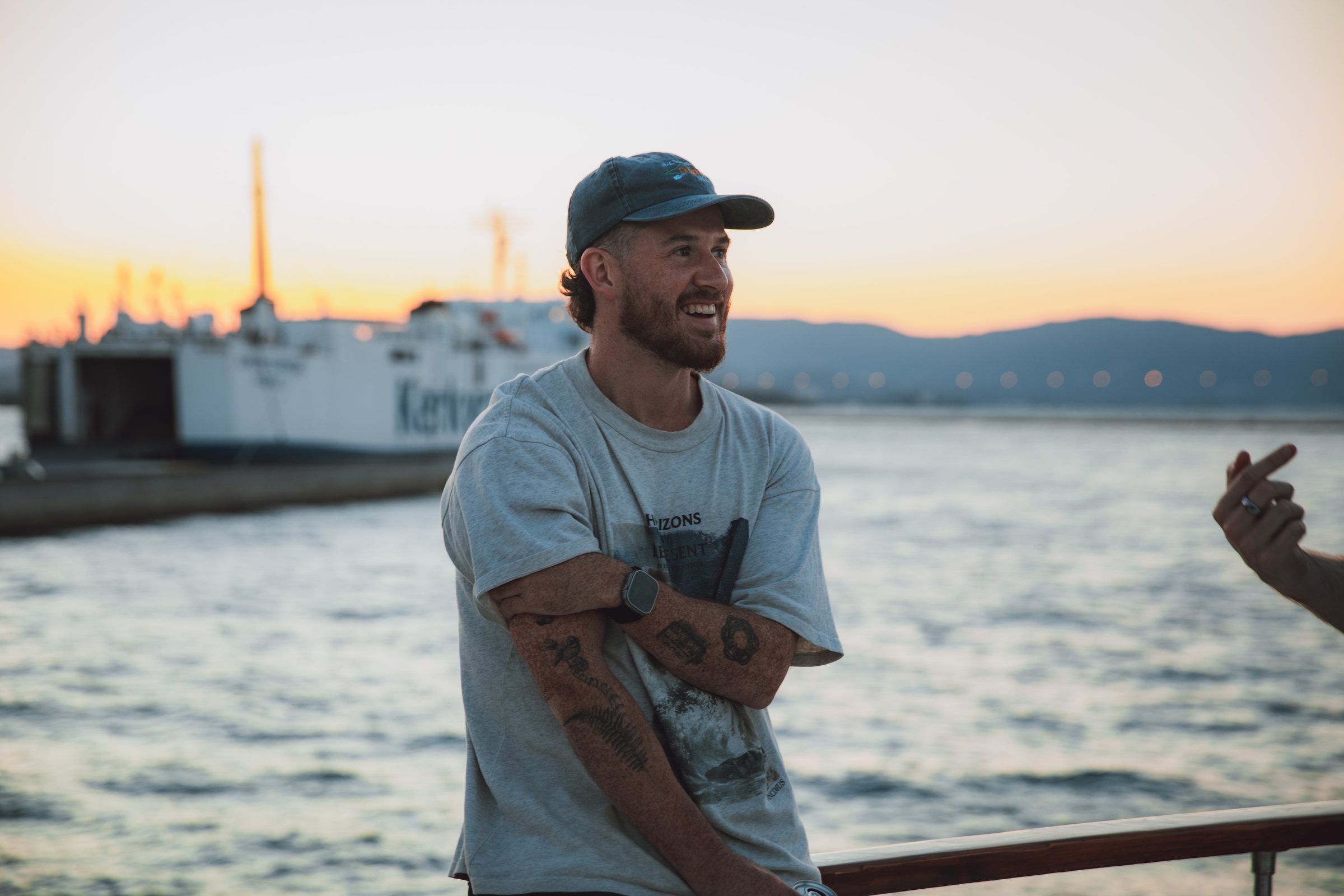 Taylor laughing on deck at sunset, the harbour and open water behind him.