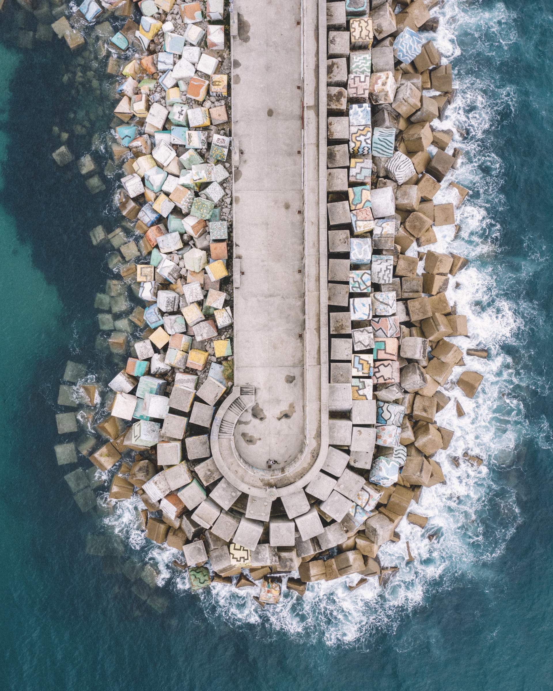 The curved end of a breakwater from above, painted blocks and white water.