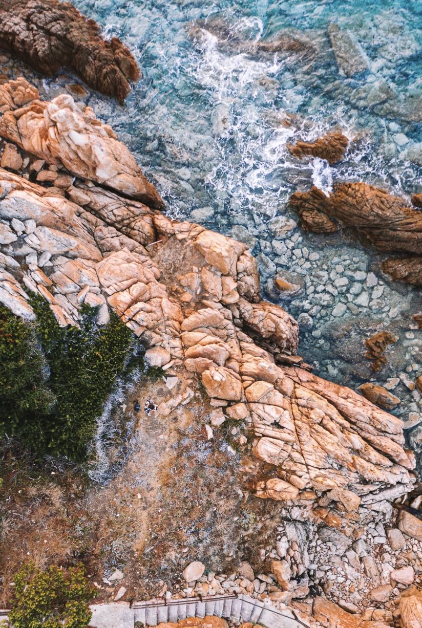 Warm granite shelf meeting turquoise surf from above, one tiny figure on the rocks.