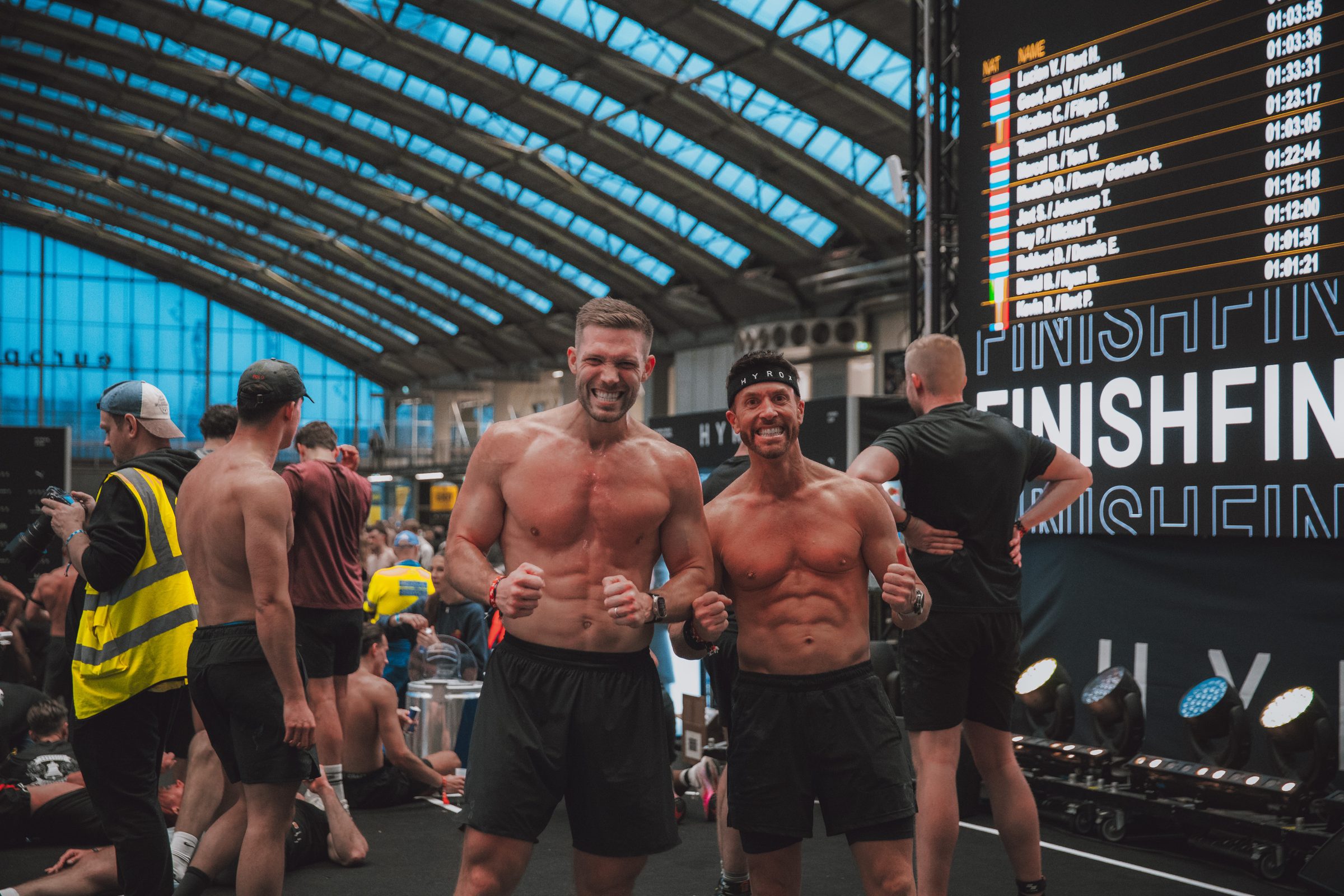 Two athletes flexing and laughing under the finish board, times still climbing.