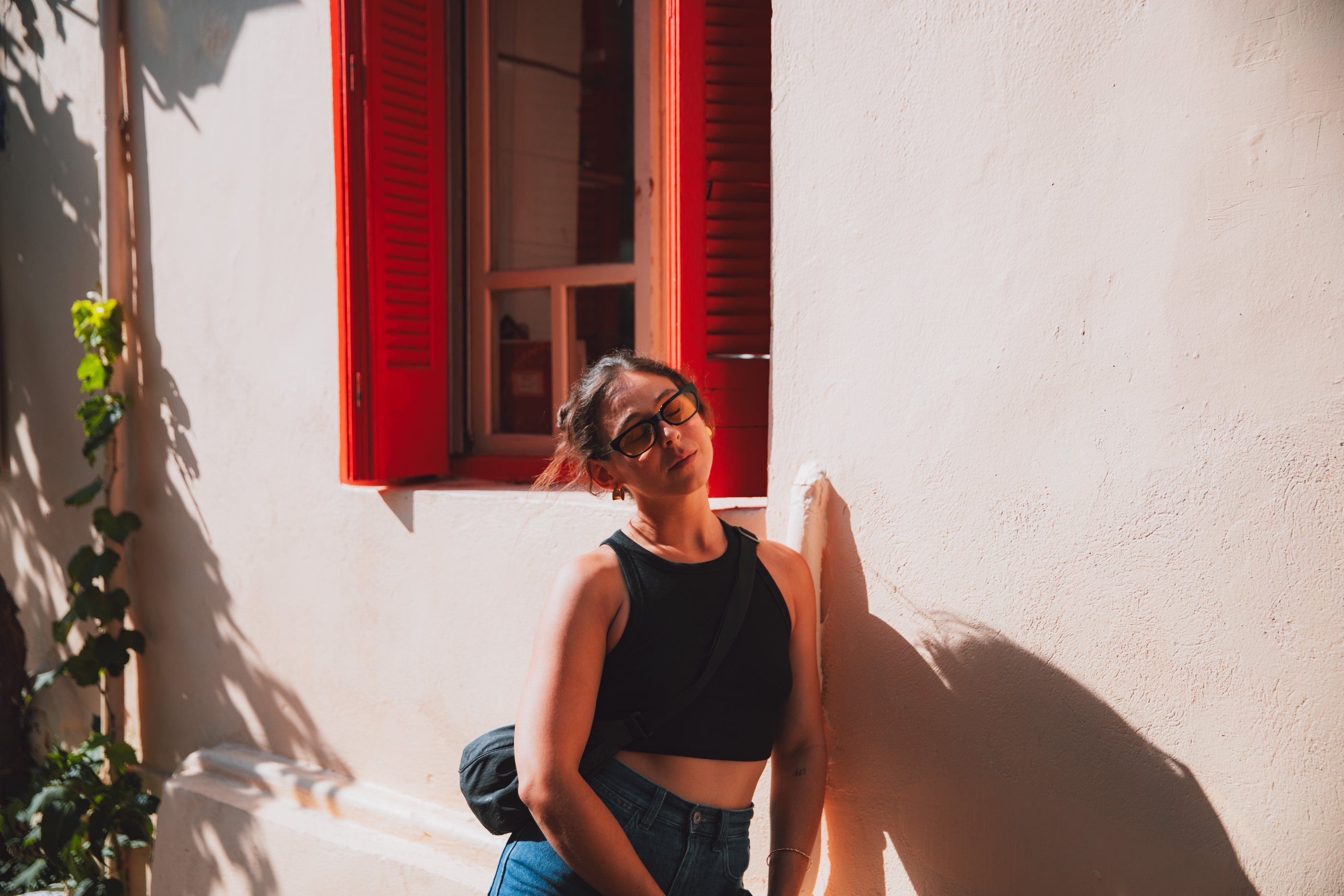 Leaning against a sunlit white wall beneath red shutters in Athens, hard shadow cutting across the frame.