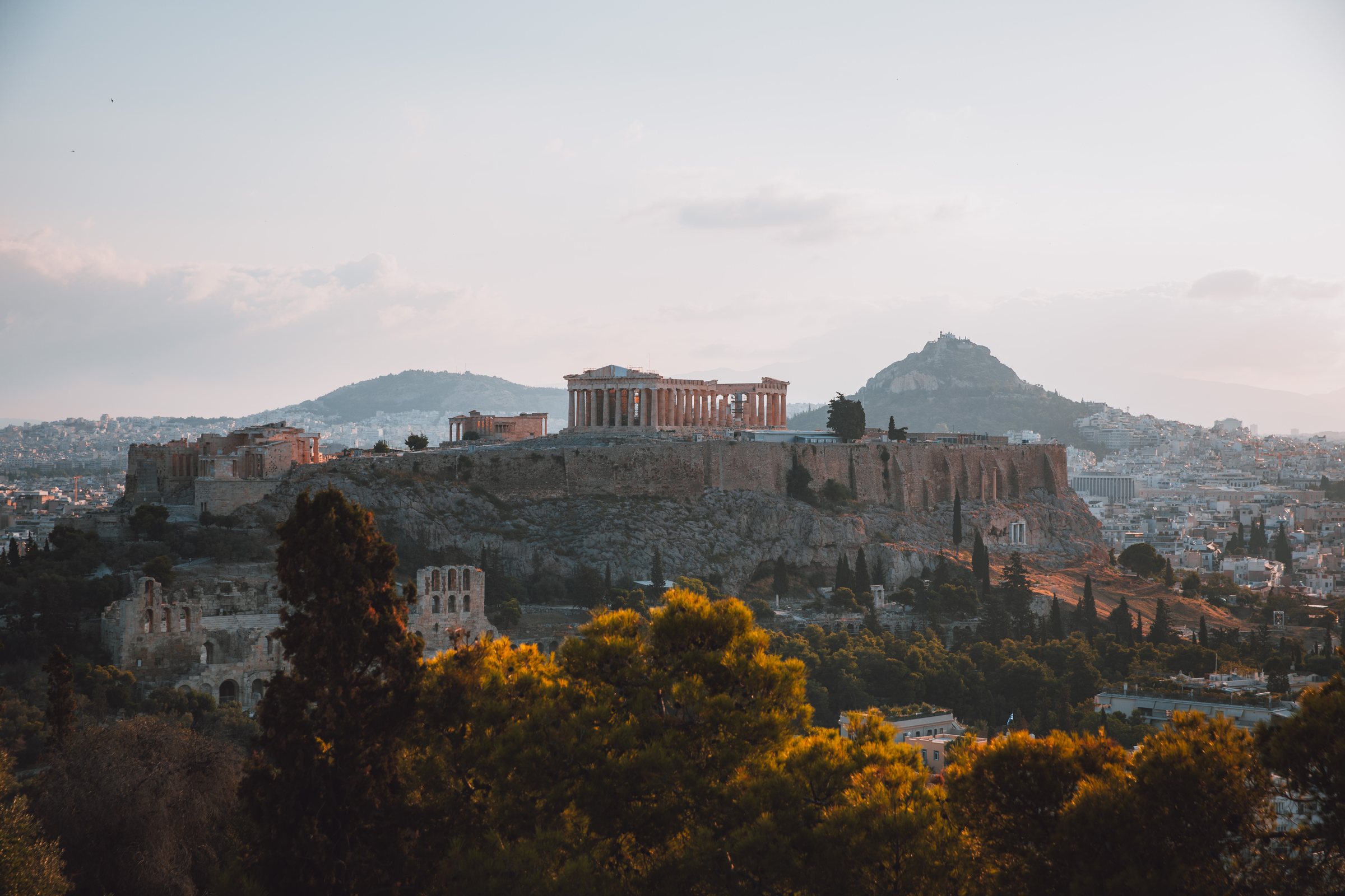 The Acropolis catching the last warm light above Athens, Lycabettus Hill beyond, pines in the foreground.